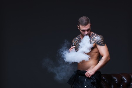 Vaper. The man with a muscular naked torso with tattoos smoke an electronic cigarette on the dark backgroundの写真素材