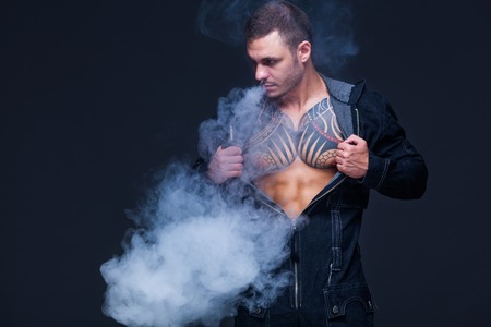 Vaper. The man with a muscular naked torso with tattoos smoke an electronic cigarette on the dark backgroundの写真素材