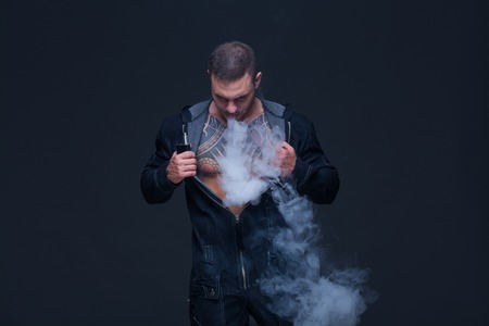 Vaper. The man with a muscular naked torso with tattoos smoke an electronic cigarette on the dark backgroundの写真素材