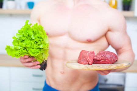 Muscular man with a naked torso in the kitchen with a salad and a piece of beef meat, the concept of a healthy diet. Athletic way of life.の写真素材