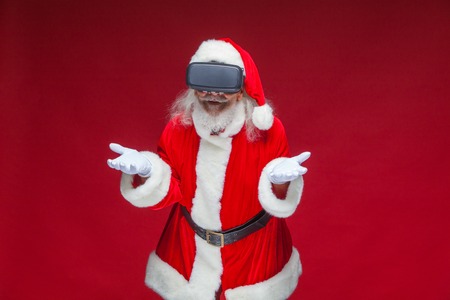 Christmas. Santa Claus in black virtual reality glasses makes gestures with his hands. Surprise, emotion. New technology. Isolated on red background.の写真素材