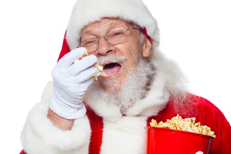 Christmas. Smiling, kind Santa Claus in white gloves with his mouth open holds a red bucket with popcorn with one hand, takes some popcorn and prepares to eat it with the second hand. The concept of visiting the cinema, watching a movie with popcorn.の写真素材