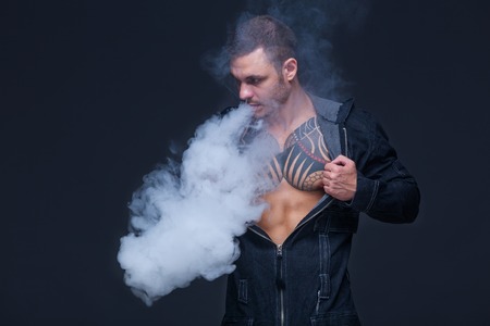 Vaper. The man with a muscular naked torso with tattoos smoke an electronic cigarette on the dark...