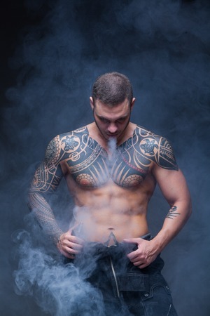 Vaper. The man with a muscular naked torso with tattoos smoke an electronic cigarette on the dark backgroundの写真素材