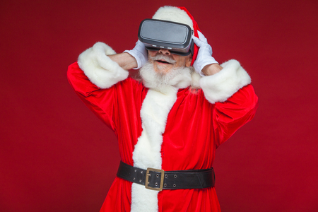 Christmas. Santa Claus in black virtual reality glasses makes gestures with his hands. Surprise, emotion. New technology. Isolated on red background.の写真素材