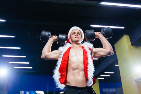Holidays and celebrations, New year, Christmas, sports, bodybuilding, healthy lifestyle - Muscular handsome sexy Santa Claus. Muscular Bodybuilder Guy Doing Exercises With Dumbbells Over in the gymの写真素材