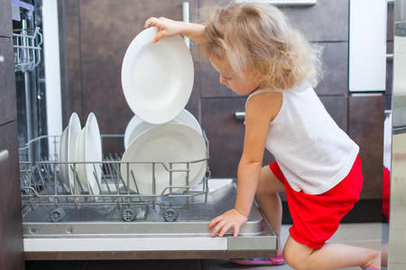 Cute blonde toddler girl helping in the kitchen taking plates out of dish washing machineの写真素材