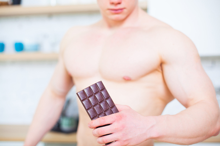 Muscular man with a naked torso in the kitchen with a tile of chocolate, the concept of a healthy diet. Athletic way of life. The temptation to sweet.の写真素材