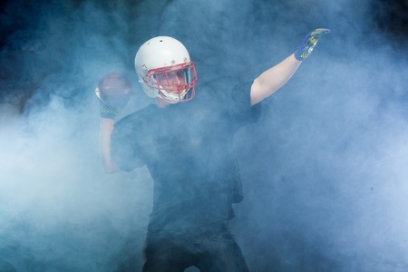 American football player in a black uniform, wearing helmet and throwing ball against white smokeの写真素材