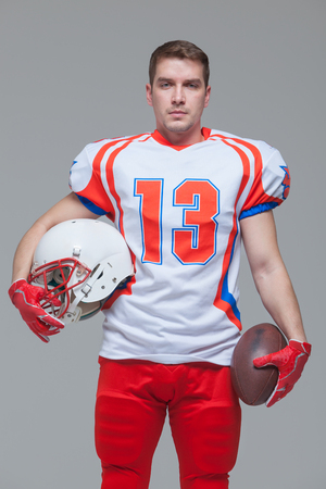 American football player holding rugby ball and helmet against grey background front viewの写真素材