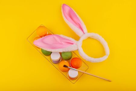 Colorful handmade Easter eggs, rabbit ears to play with, and jars of multicolored gouache with tassels.. Top view of an open transparent box. Isolated on a yellow background. Preparing for Easterの写真素材