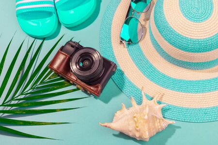 Top view on beach accessories on turquoise blue background - sunglasses, blue flip-flops, striped hat, camera, shell and palm leaf. Concept of the long-awaited vacation at sea and travel. Advertising spaceの写真素材