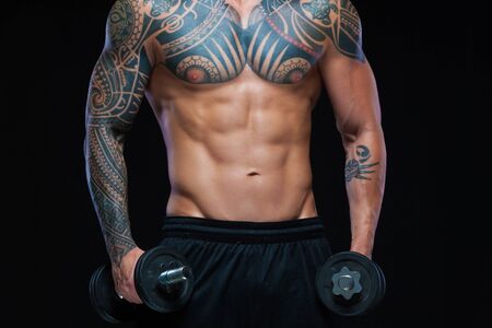 Close up of a tattooed muscular man torso holding dumbbells on black backgroundの写真素材