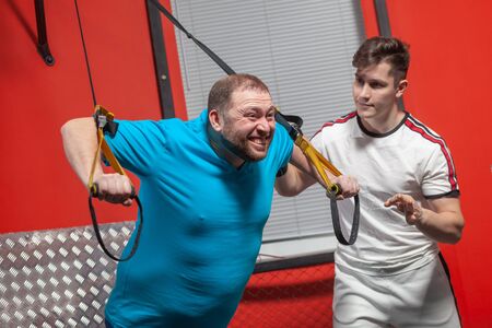 Fat man in a gym is diligently doing pull-ups under the guidance of a personal trainer.の写真素材