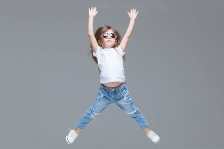 Kid girl preschooler in blue jeans, white t-shirt and sunglasses is jumping with raised hands isolated on grey backgroundの写真素材