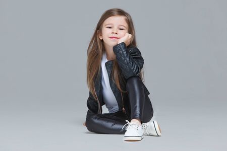 Baby girl with long hair in black leather jacket and leggings is sitting in the studio on grey backgroundの写真素材