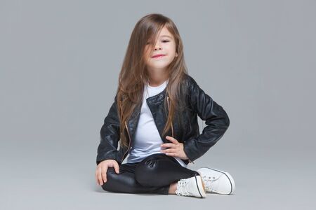 Baby girl with long hair in black leather jacket and leggings is sitting in the studio on grey backgroundの写真素材