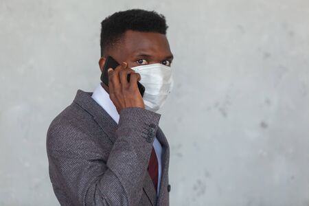 COVID-19. Handsome African black man in a suit using medical mask as protection from coronavirus pandemic talking on a mobile phone against a gray concrete wallの写真素材
