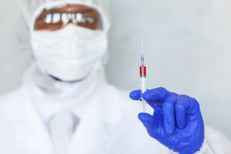 Doctor in chemical protection suit is holding syringe with blood sample fot testing it on COVID, COVID-19, coronavirus to stop pandemic and epidemic of the virusの写真素材