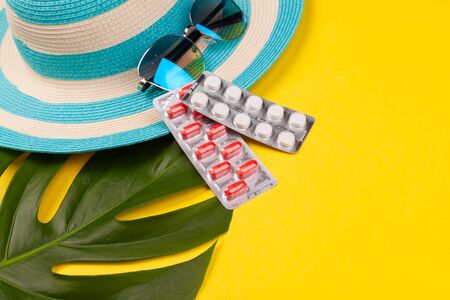 Top view on white and red pills in blister pack, blue hat, sunglasses, monstera leaf on yellow background. Concept of beach holiday, sea tour, warm sunny summer. Acclimatization, illness during travel, health insurance. Advertising space. COVID, COVID-19, coronavirus, pandemic, epidemic, virus,の写真素材
