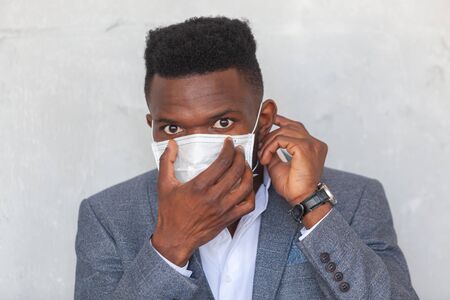 COVID-19. Young African man puts on a protective medical face mask to prevent infection with coronavirus and pandemic standing in a suit against a gray concrete wallの写真素材
