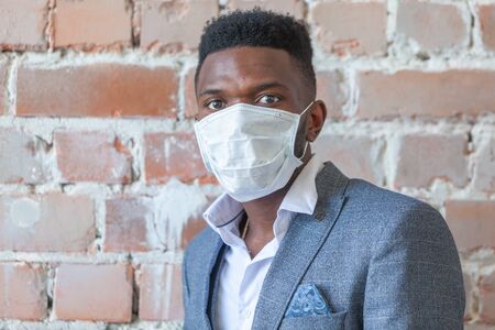 Close up of young African black man in a suit against a red brick wall is afraid of a coronavirus epidemic and wears a medical mask so as not to get infected COVID-19の写真素材
