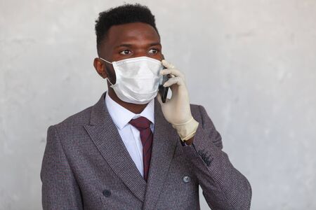 COVID-19. Handsome African black man in a suit is using medical mask and holding his mobile phone with a hand in gloves. Measures to prevent coronavirus infection and the development of a pandemicの写真素材