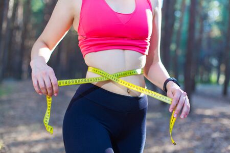 Photo of a fit and healthy young lady measuring her waist with a tape measure controlling her weight loss while training in the forest.の写真素材