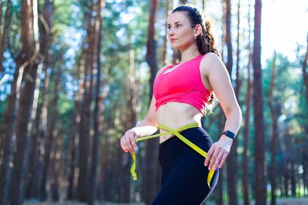 Photo of a fit and healthy young lady measuring her waist with a tape measure controlling her weight loss while training in the forestの写真素材