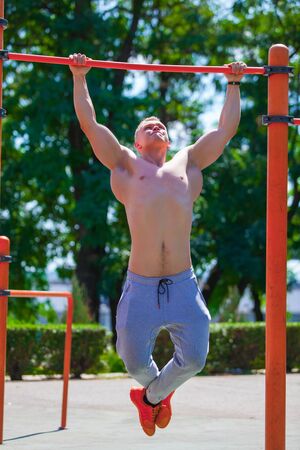 Strong muscular man with a bare torso pulls himself up on a crossbar. Working out outdoors in summer...