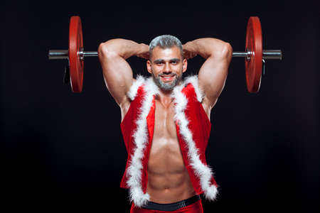 Sexy Santa Claus. Bodybuilder young handsome santa clause smile train muscles arms and triceps at New Years eve and Christmas winter holiday black background.の写真素材