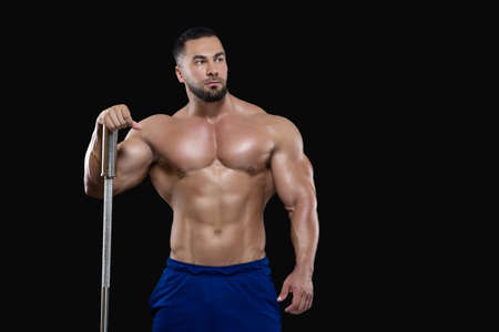 Young sexy athlete man is standing leaning with one hand on the barbell isolated on black background