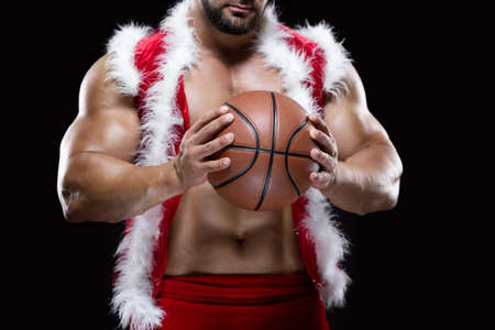 Front view portrait of an attractive muscular Santa Claus holding a basketball with both hands isolated on black backgroundの写真素材