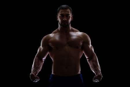 Portrait of a bodybuilder standing isolated on black background in a shadow with clenched fists to show off his musclesの写真素材
