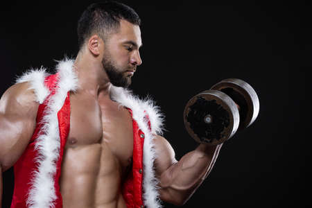Strong muscular athlete wearing Santa Claus costume over naked body is lifting a dumbbell with one hand isolated on black backgroundの写真素材