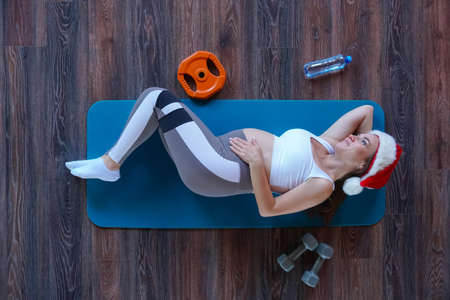 Top view of young beautiful pregnant girl in Santa Claus hat lying on the mat on the floor in a gym near to dumbbells, a water bottle and a discの写真素材