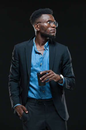 Wealthy and serious dark-skinned businessman is standing with a cup of coffee isolated on black backgroundの写真素材