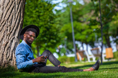 Portrait of a young black man in a hat and glasses sitting under tree in the park holding a cup of coffee in one hand and a mobile phone in the otherの写真素材