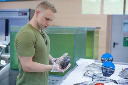 Young male athlete in green T-shirt and gloves is holding frozen fish choosing food in a grocery...