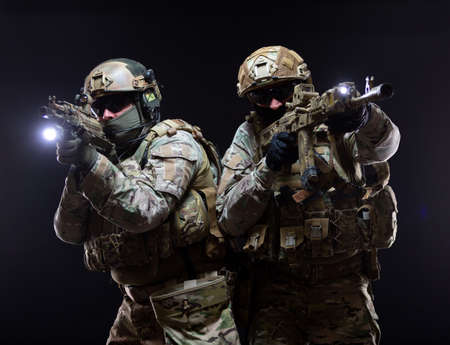Two soldiers in military gear and bulletproof vests cover each other and raise their submachine guns to their faces taking aim, in full combat readiness, isolated on black backgroundの写真素材