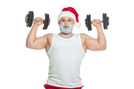 A big athletic male athlete trains biceps lifting dumbbells standing in a Santa Claus hat ready to celebrate Christmas isolated on a white backgroundの写真素材