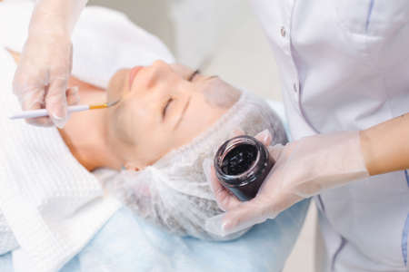 Hands of cosmetology specialist applying carbon nanogel on young woman patient face using brush,...