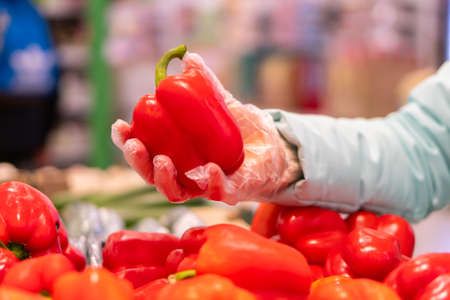 Closeup of woman hands in gloves choosing red bell peppers in a supermarket. Shopping during quarantine in compliance with the rules to prevent the spread of coronavirus infection in a pandemicの写真素材