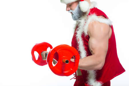Christmas. Strong muscular Santa Claus with grey beard wearing christmas hat and red shorts is lifting a heavy barbell in a gym performing a workout isolated on white background.の写真素材