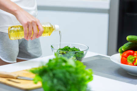 Close-up photo of a girl in shorts pouring vegetable oil on a vegetable vitamin salad in a glass bowl in the kitchenの写真素材