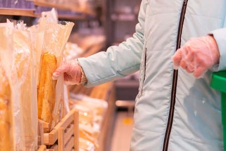 Close up of woman hands in gloves holding a shopping basket and choosing bread. Shopping during quarantine in compliance with the rules to prevent the spread of coronavirus infection in a pandemicの写真素材