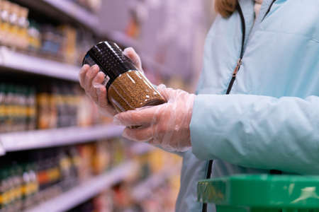 Close up of woman hands in gloves holding a shopping basket and choosing coffee in a supermarket. Shopping during quarantine in compliance with the rules to prevent the spread of coronavirus infection in a pandemic.の写真素材