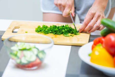 Close up of a girl hands cutting greenery with a knife on a cutting board for a vegan vitamin vegetable salad and putting them in a glass bowl while cooking breakfast in the kitchenの写真素材