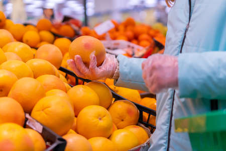 Photo of a cute girl in a medical mask and gloves choosing tangerines in the supermarket. Shopping in compliance with anti-epidemic measures during the coronavirus pandemicの写真素材