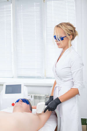 A pretty cosmetologist and her client in blue safety glasses during the procedure of laser hair removal from the armpit of a young manの写真素材